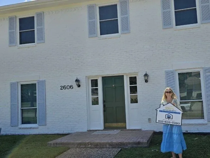 Happy homeowner holding a 100 percent satisfied sign in front of a freshly painted white brick home with blue shutters