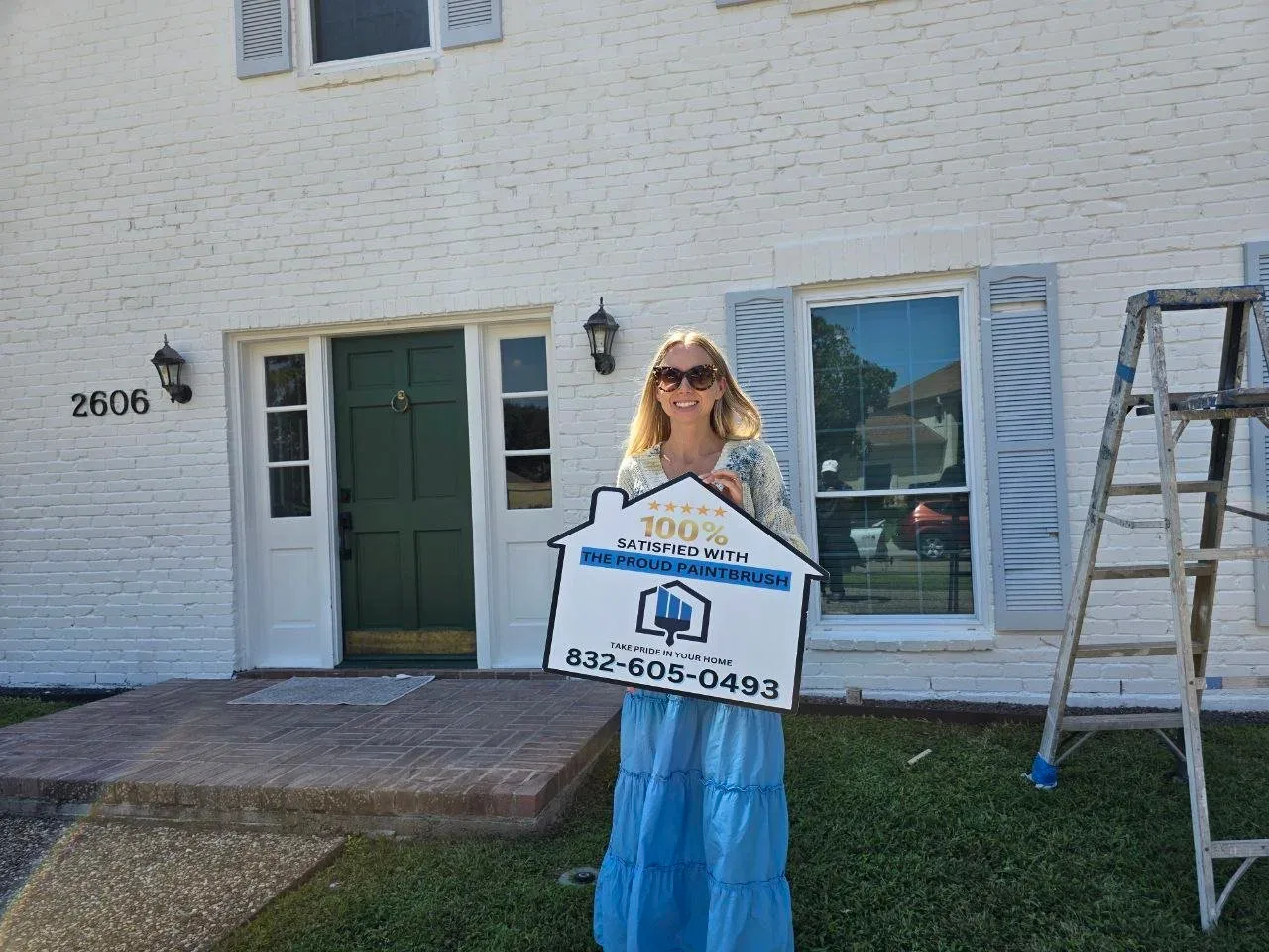 Woman standing outside a white brick house holding a sign for a painting business, with a ladder to her right, smiling and wearing sunglasses.